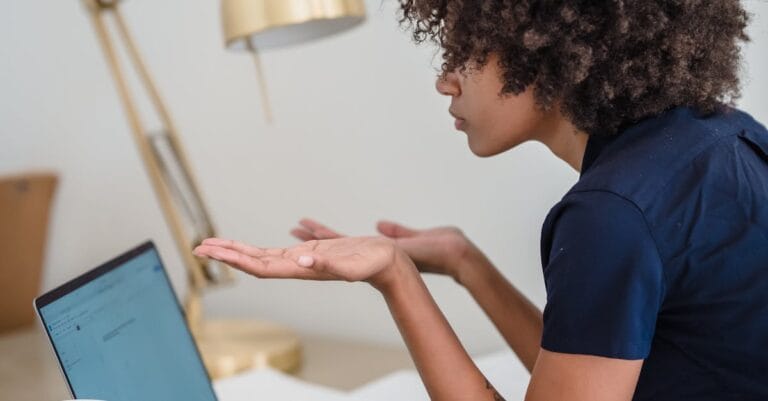 Woman Sitting Behind a Desk, Using Laptop and Spreading Her Hands as a Sign of Not Understanding Something